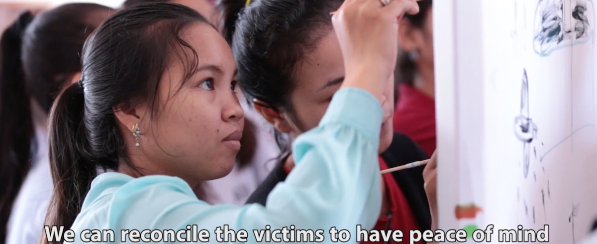 Screenshot of a video where a girl is drawing on a board. The caption says "We can reconcile the victims to have peace of mind and the communities to understand more."