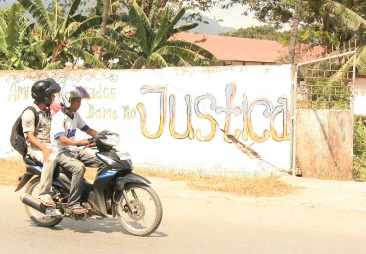 Two people on a motorcycle look at a mural next to them that says "Justica."