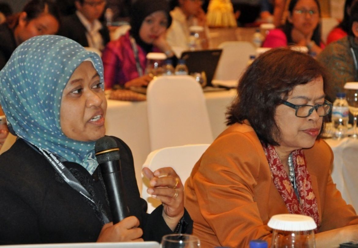 Two women seated at tables in a conference hall, one with a microphone