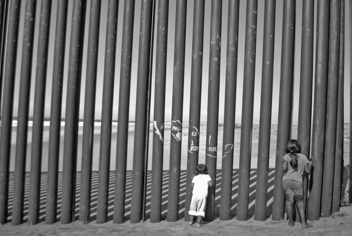 Black and white image of children facing away from camera looking through a border fence